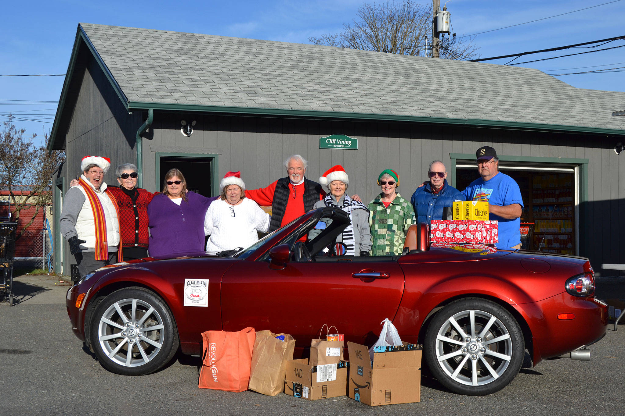 Olympic Peninsula Chapter of Club Miata Northwest members gather with Stephen Rosales, board president of the Sequim Food Bank, around Vicki Burr-Chellin’s 2008 Miata MX5 for a donation on Dec. 14 of $350 and several boxes of food. Sequim Gazette photo by Matthew Nash