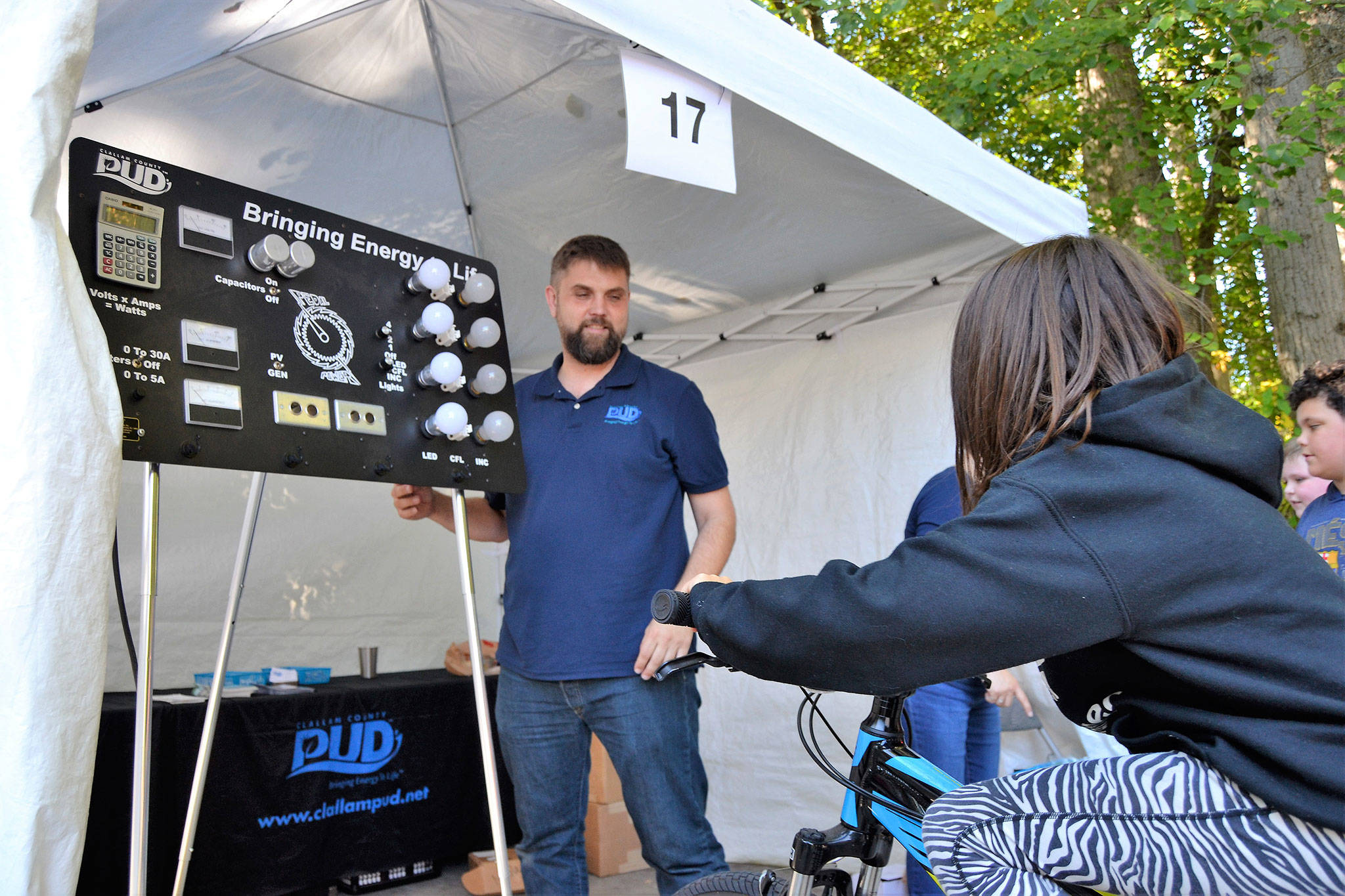 Olivia Nelson, 9, rides a bike to see how much energy it takes to light different bulbs with help from Mattias Järvegren, utilities services coordinator for Clallam PUD. Sequim Gazette photo by Matthew Nash