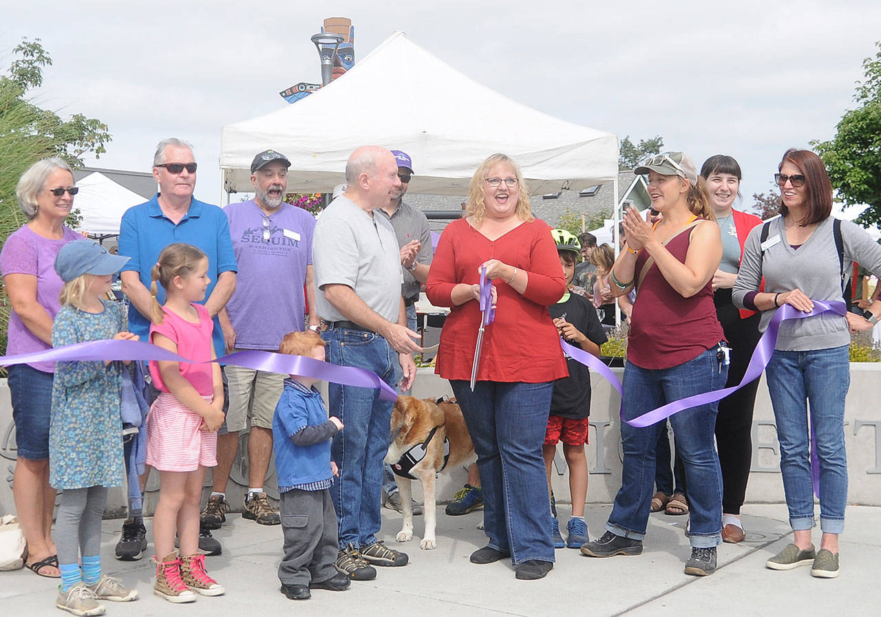 Gina Witz, Sequim Farmers Market board president (at center), cuts a ceremonial ribbon on at the market on Aug. 17, celebrating the market’s designation as a nonprofit organization. Joining her are, among other dignitaries, Sequim Mayor Dennis Smith (center left). Sequim Gazette photo by Michael Dashiell