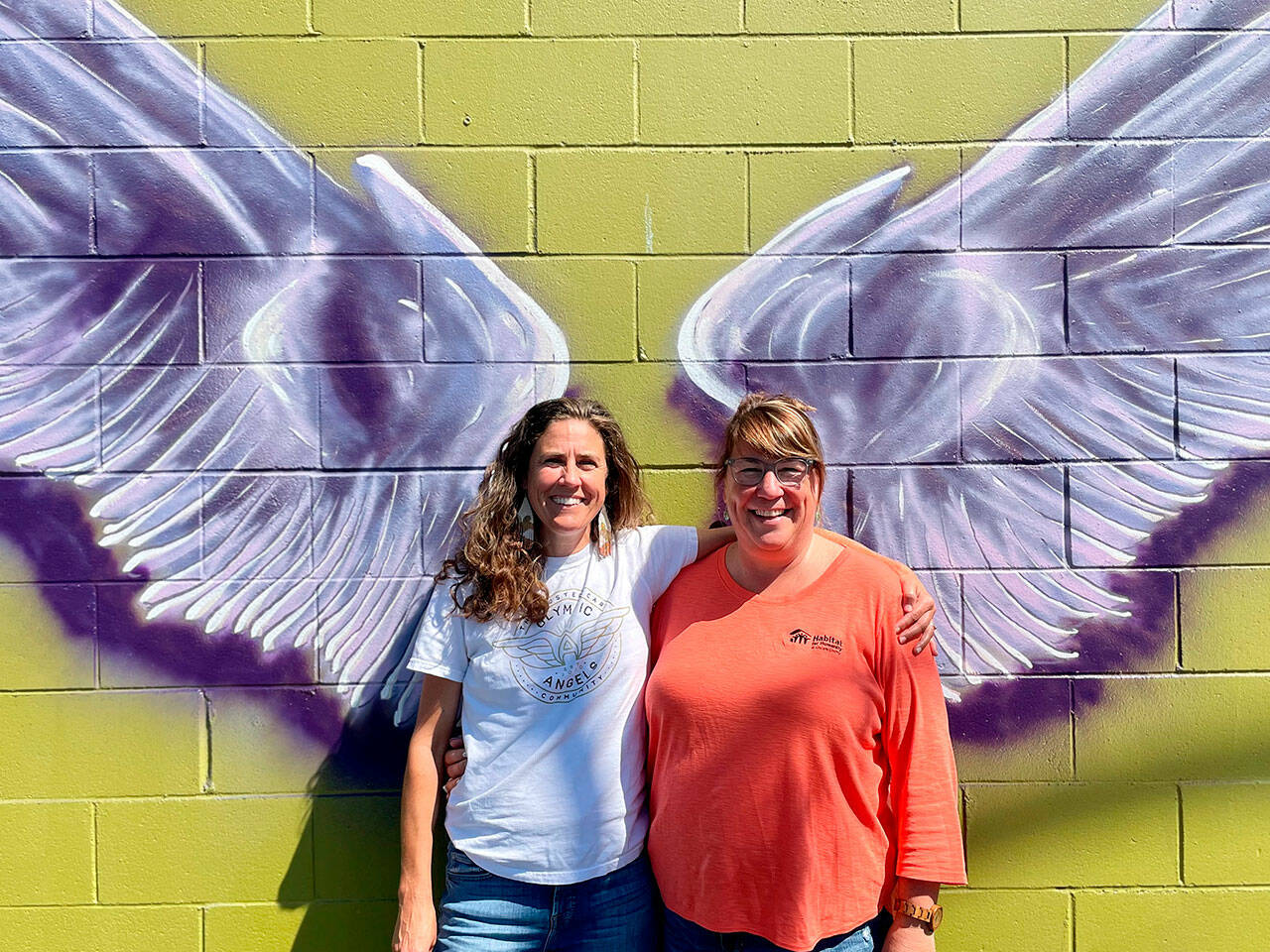 Sequim Gazette photo by Matthew Nash/ A new mural by Craig Robinson in downtown Sequim represents a new partnership between Olympic Angels and Habitat for Humanity of Clallam County. Executive director Morgan Hanna with Olympic Angels, on left, said they seek volunteers and mentors to help foster families. Colleen Robinson, chief executive officer for Habitat, said people are welcome to take photos on the wall of Habitat’s Boutique Store and they’ll place a sandwich board sign with information outside, and provide pamphlets about Olympic Angels during business hours.