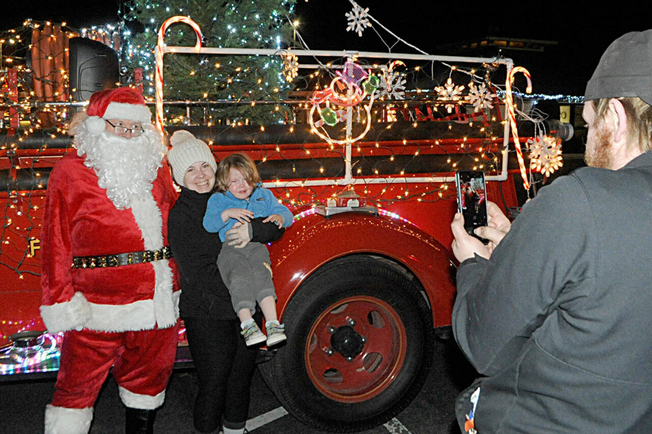 Sequim Gazette file photo by Matthew Nash/ Veronica Weatherly holds her son Tucker for a photo-op with Santa Claus in 2021 as her husband Doug takes a photo. Santa’s Toy and Food Fire Brigade tours Sequim again Dec. 4-7.