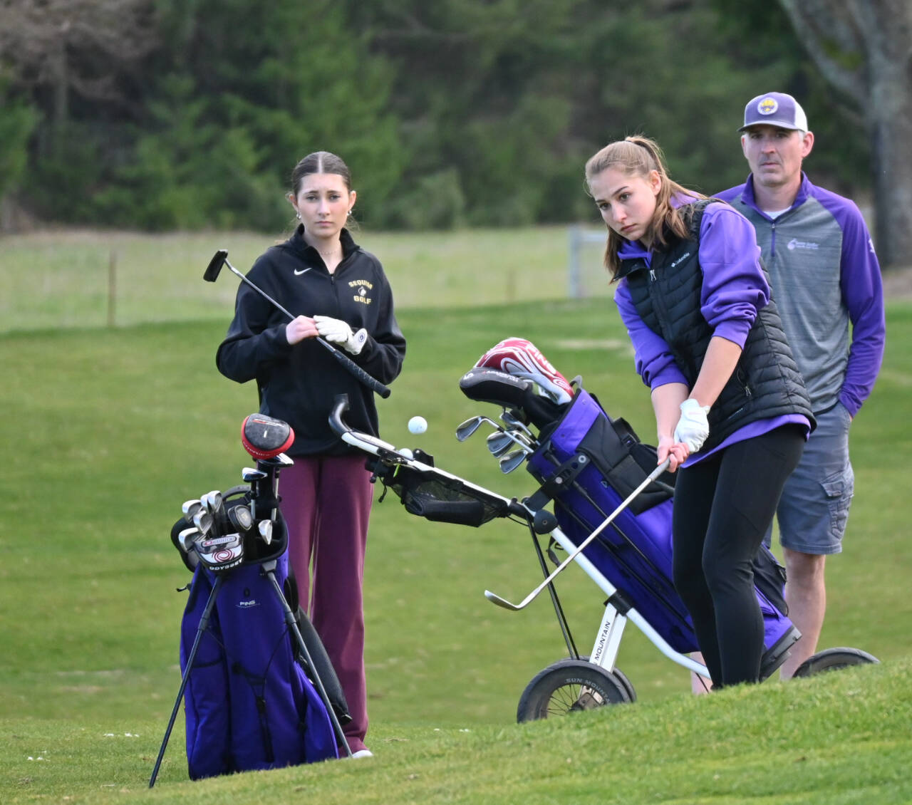 Sequim Gazette photo by Michael Dashiell
With teammate Kendra Dodson and assistant coach Darren Stephens looking on, Taryn Johnson chips a shot onto the green on the third hole at The Cedars at Dungeness in a March 21 match against Bainbridge.