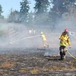 Fire District 3 personnel work on putting out flames that are melting the plastic siding from a shed in a brush fire in Carlsborg on July 26. Sequim Gazette photo by Conor Dowley.