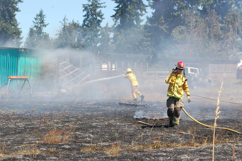Fire District 3 personnel work on putting out flames that are melting the plastic siding from a shed in a brush fire in Carlsborg on July 26. Sequim Gazette photo by Conor Dowley.