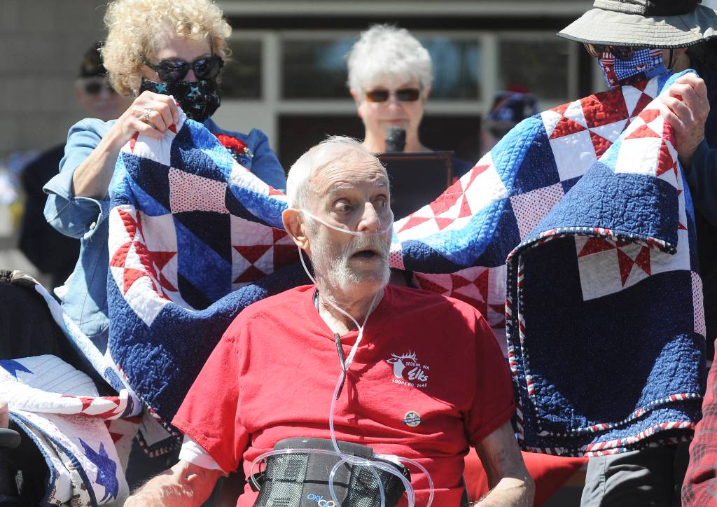 Above: With Kathryn Bates  team leader of the North Olympic Peninsula Quilts of Valor group  looking on, volunteers drape a patriotic quilt on Frank Hurlbut during a special Memorial Day ceremony at Sinclair Place on May 29. Sequim Gazette photos by Michael Dashiell
At left: Betsy Schultz speaks at a special memorial Day event at Sinclair Place on May 29. Schultz, whose Captain Joseph House lends support to families dealing with the death of a family member in the services, said Saturday was the 10th anniversary of her sons passing.