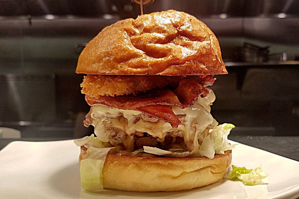 The Cowboy features three patties with Tillamook pepper jack cheese, applewood smoked bacon, house BBQ sauce, chopped onions, pickles, an onion ring, lettuce, and tomato on an artisan bun. Photo courtesy of Shadowline Burgers and Brews