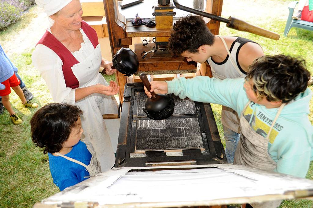 Nickie Allen of Utah shows brothers Rizzolo, 9, Canaan, 12, and Broden Texeira, 17 of Bremerton how her husband Goves replica of Isaiah Thomas printing press works. The Allens returned to the Northwest Colonial Festival for the first time since 2017 to share hands-on learning.