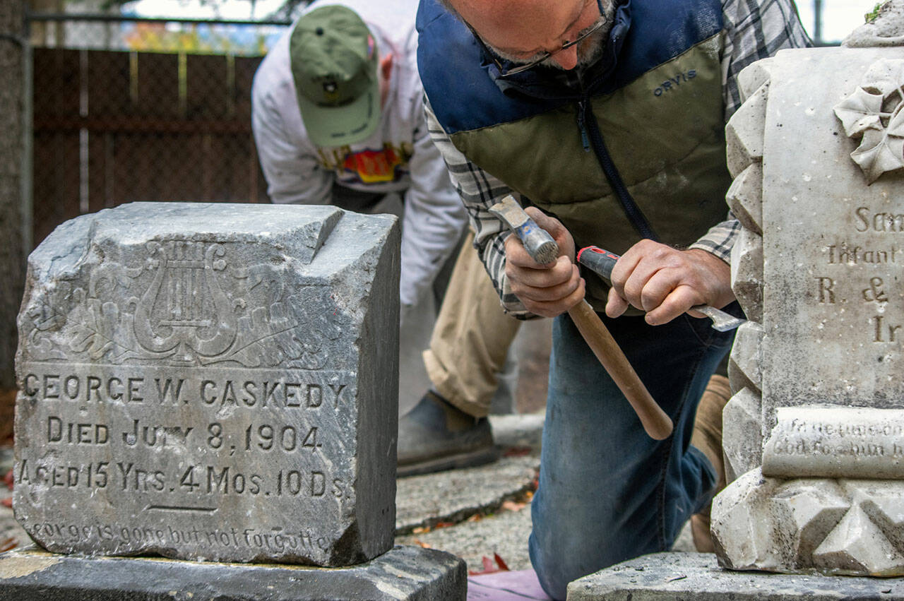 Using a chisel and hammer, Chris Smith chips away white mortar from the headstone of the infant son of R. and Eleanor Irwin, "God for him his angels sent," can be read. The inscription will become gradually more legible from being sprayed by volunteers with D/2 Biological Solution (TM). Next to the infant's headstone, sits that of George W. Caskedy. "Not a lot of information is available on Caskedy," said Priscilla Hudson, Sequim Prairie Garden Club historian, "because he was such a young kid. His father was W.M. Caskedy, born in Ohio. His mother Emma was born in Minnesota." Caskedy was fifteen when he died. Hudson said that there was no death certificate or newspaper reference, and nothing at the LDS family history center. "I'm assuming the father did logging," as they moved quite a bit, with residences in Blynn, Sequim, and Rema, all logging areas. George Caskedy's headstone was cleaned and reset in the corner of Pioneer Memorial Park with a collection of other headstones from the late 1800s to the early 1900s by volunteers this autumn. Sequim Gazette photo by Emily Matthiessen
