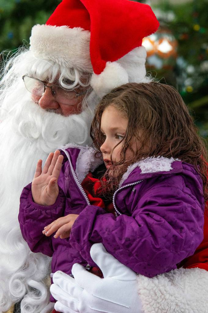 Noel Berra, age 4, waves from Sequim Santas lap at the Hometown Holidays, an usually annual event in Sequim. Although the day was gray and rainy, a spirit of good cheer was pervasive in the crowd who gathered at the Centennial Square on the corner of Washington Street and Sequim Avenue to listen to the Sequim City Orchestra play, buy hot drinks from the cub scouts and meet Santa and the Irrigation Festival Royalty. Peninsula Daily News photograph by Emily Matthiessen.