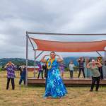 Sequim Gazette photo by Emily Matthiessen / Alakaʻi in training, Casara Desiree Hemakanamaikapu`uwai Caro leads members of a Sequim audience in a Hawaiian dance in the field behind the Sequim Library on July 16. Her mother, Candace Marie Kananiokamokihana Melendez of Hālau Hula Ka Lei Mokihana I Ka Ua Noe, plays the ukulele on the stage. North Olympic Library System hosted the dance, history and language workshop as part of their summer reading program.