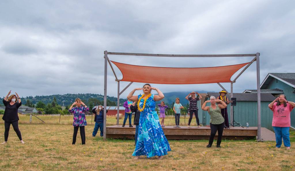 Sequim Gazette photo by Emily Matthiessen / Alakaʻi in training, Casara Desiree Hemakanamaikapu`uwai Caro leads members of a Sequim audience in a Hawaiian dance in the field behind the Sequim Library on July 16. Her mother, Candace Marie Kananiokamokihana Melendez of Hālau Hula Ka Lei Mokihana I Ka Ua Noe, plays the ukulele on the stage. North Olympic Library System hosted the dance, history and language workshop as part of their summer reading program.
