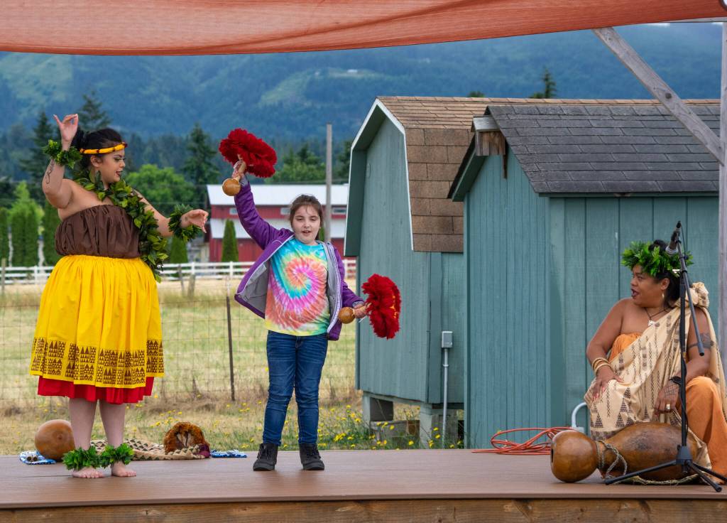 Sequim Gazette photo by Emily Matthiessen
Kumu Hula Mokihana Melendez (left) and featured dancer Alakai Makana Caro (right) of Halau Hula Ka Lei Mokihana I Ka Ua Noe invite Sequim resident Amaya Franich to try the uliuli, a type of gourd rattle with stones or seeds and feathers. Franich, participating in this NOLS summer reading event, said it was fun.