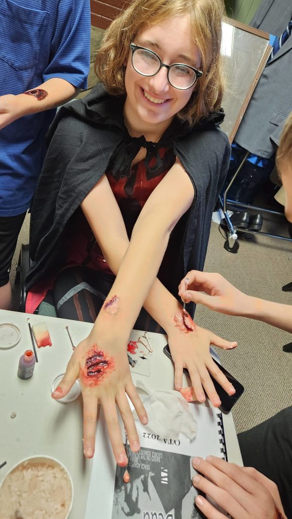 Photo courtesy of Karlene Minea / Georgia Bullard beams as make-up is applied to her hands and arms during a rehearsal for Night of the Living Dead, Olympic Theatre Arts' upcoming play.
