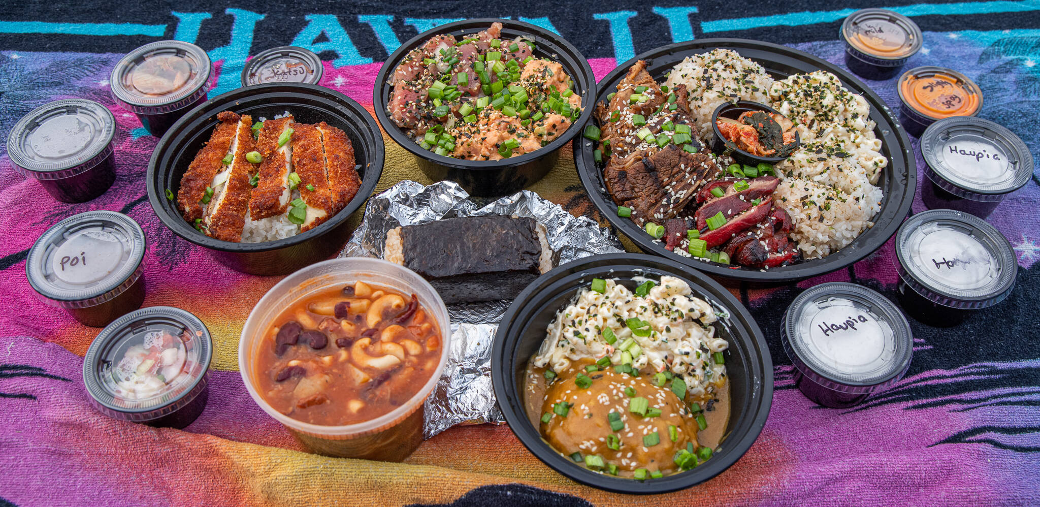 Sequim Gazette photo by Emily Matthiessen / Take out from Da Kindz Island Grill brings new flavors and smells to a chilly Pacific Northwest Day. The ring of big bowls, clockwise from top center: poke bowl with half spicy and half shoyu ahe and rice; barbacue mix plate with short ribs; chicken and char sui port; with rice and kim chee; vegetarian curry and rice with macaroni salad; Portuguese bean soup, and chicken katsu for keiki (children). In the middle is a portion of spam musubi, and surrounding the entrees are sides like haupia (coconut pudding), poi, kim chee and sauces.