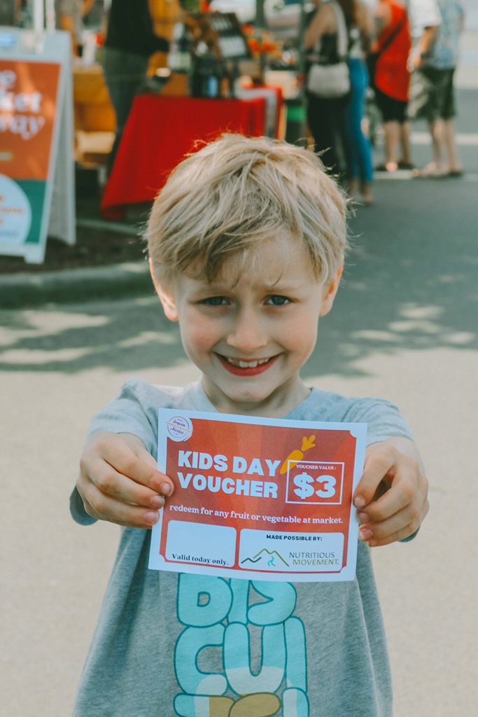 Photo courtesy Sequim Farmers & Artisans Market/ Ollie, a market guest, holds up a Kids Day voucher. Thanks to Nutritious Movements partnership, were able to facilitate hands-on experience with local food for all ages.