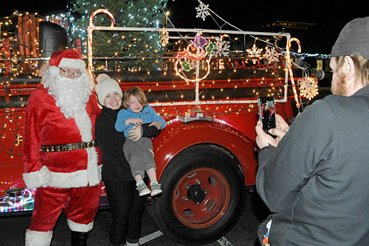Sequim Gazette file photo by Matthew Nash/ Veronica Weatherly holds her son Tucker for a photo-op with Santa Claus in 2021 as her husband Doug takes a photo. Santas Toy and Food Fire Brigade tours Sequim again Dec. 4-7.