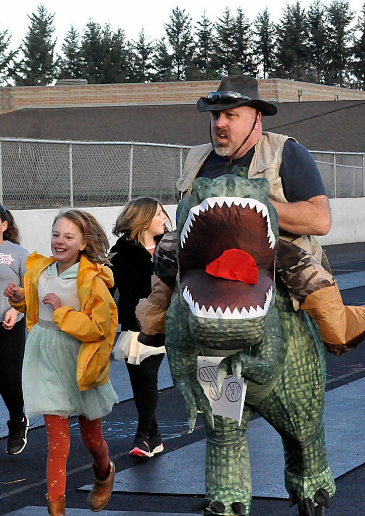At right: Third-grader Dixie Utz, teacher Greg Newton and Tom the dinosaur run along the Sequim School District track during the Turkey Trot.