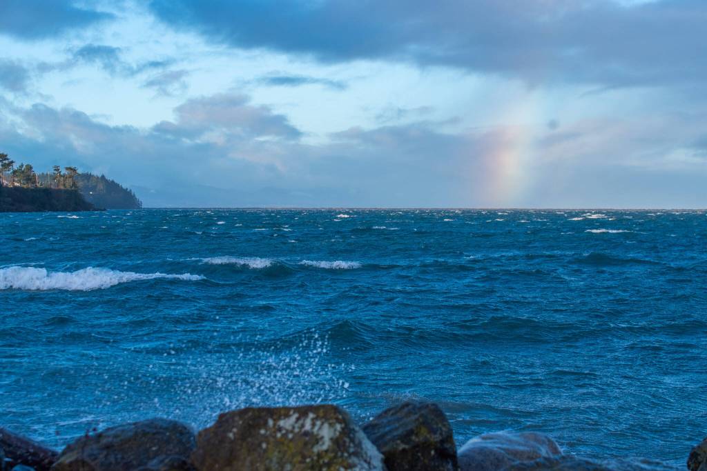 Sequim Gazette photo by Emily Matthiessen/ High waves at Cline Spit on Fri. Jan 10 splash into the parking lot while a brief storm brings a chunk of rainbow over the sea.