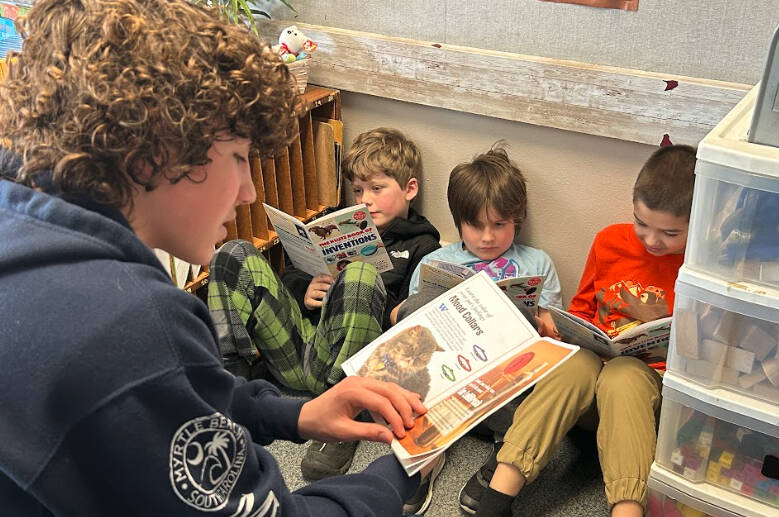 Sequim Gazette photo by Matthew Nash/ Sequim eighth grader Silas Hagar reads about inventions with Greywolf second graders, second from left, Calvin Crews, Wyatt Hamilton, and Xander Furbee.