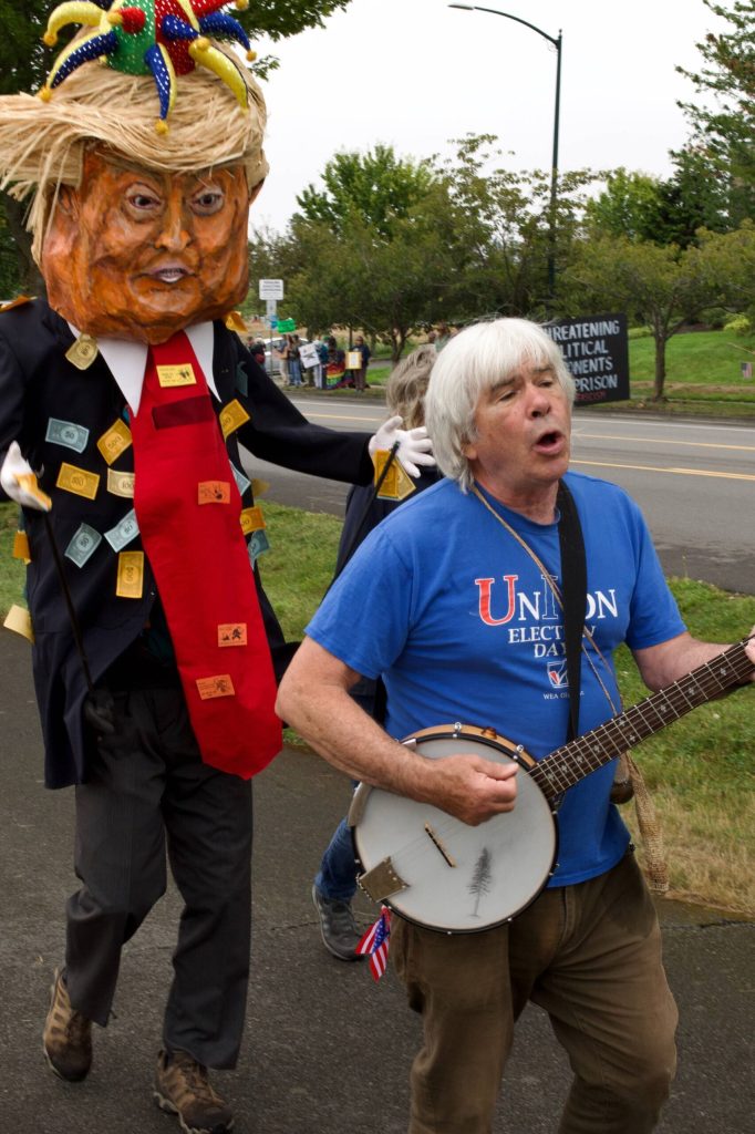 Steve Koehler of Sequim plays his banjo and sings This Land is Your Land with a caricature of Trump walking behind him.