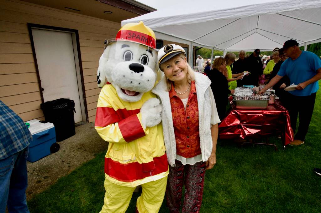 Event organizer Becky Penrose poses with Clallam County Fire District 3's  Sparky the Fire Dog as attendees of Saturday's community picnic enjoy burgers from Sequim's Black Bear Diner.