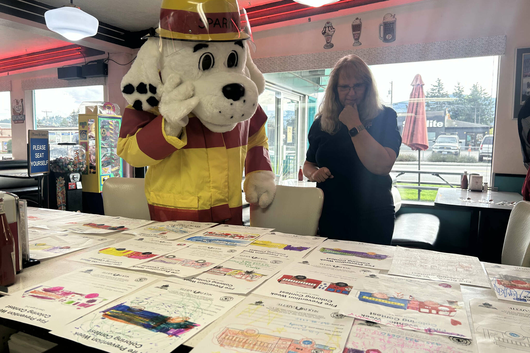 Sequim Gazette photo by Kathy Cruz/
Sparky the Fire Dog carefully considers the Fire Prevention Week contest entries laid out on the counter at Hi-Way 101 Diner by Clallam County Fire District 3 volunteer EMT Lisa Law.