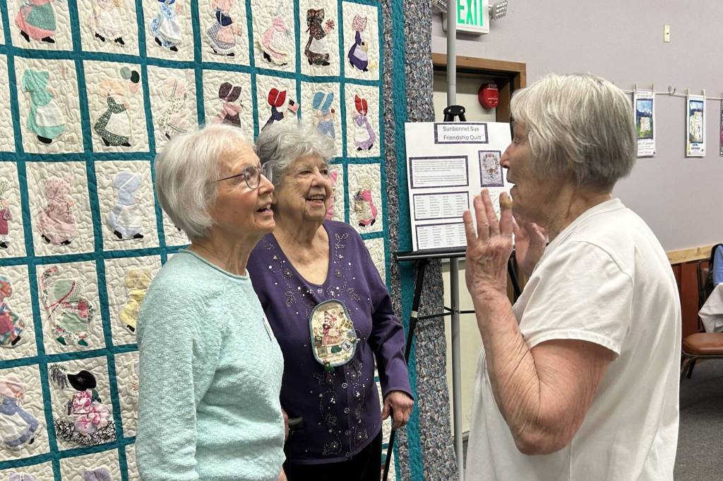 Sequim Gazette photo by Matthew Nash/ 
Longtime Sunbonnet Sue Quilt Club members, from left, Nancy Davis, Alicia Crawford, and Joanne Thoma share a story during the club’s 40th anniversary party. They’re standing by the “Sunbonnet Sue Friendship Quilt” that was one of a handful of quilts on display at the event.