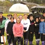 Olympic Game Farms hybrid softball team Chaos Kittens stand under umbrellas as they await Sequim Little Leagues opening day ceremonies. The girls started the season with machine pitching and will transition to player pitching as the season progresses.