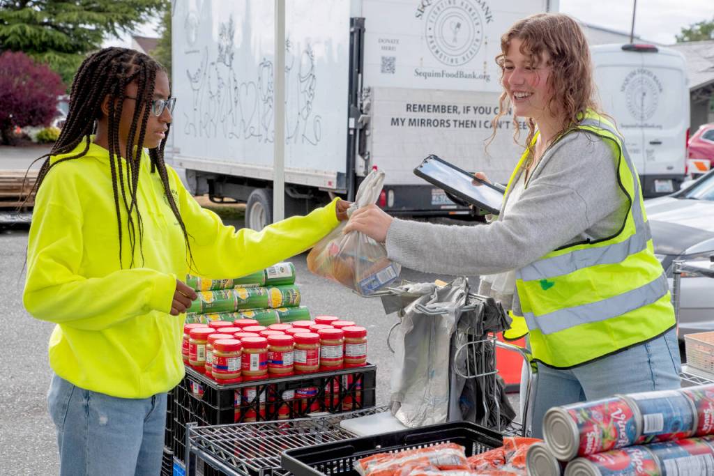 Longtime Sequim Food Bank helper Skylar Kryzworz, who graduated from Sequim High School last year and continues to volunteer when she comes home from college, hands a plastic bag full of food items to teen volunteer Trinity McNorris.