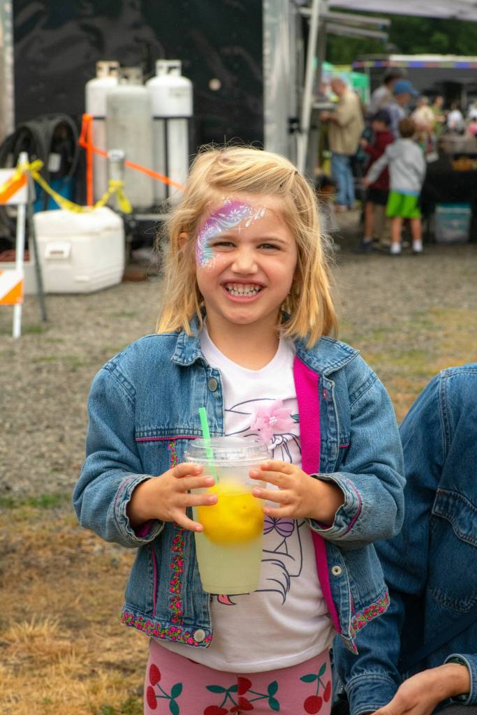 Sequim Gazette photo by Monica Berkseth/ Mabel Lepping. 4, of Sequim, is all smiles holding lemonade after getting her face painted for the first time at Frida Haas face painting studio during the Sequim Lavender Festival in the Park.
